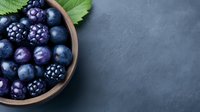 Fresh dark berries including blueberries, blackberries and plums in wooden bowl on dark slate background with green leaves and copy space for text.