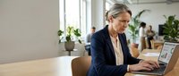 Professional mature Caucasian businesswoman working on laptop in modern office environment with colleagues in background for corporate presentations.