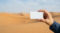 Hand holding blank white business card against sandy desert landscape with dunes and clear sky, suitable for mockup and branding projects.