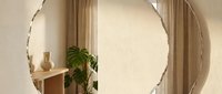 Young woman with medium skin tone, wearing casual beige clothing, standing in modern minimalist interior with round mirror, potted plant, and neutral curtains. Suitable for lifestyle, home decor, and interior design projects.