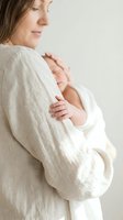 Tender moment of Caucasian mother in beige knit sweater gently holding sleeping newborn baby in soft white blanket against neutral background.