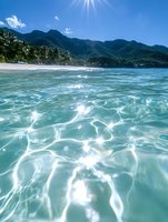 Crystal clear turquoise water with sunlight creating sparkles on surface, tropical mountain coastline and sandy beach in background on sunny summer day.