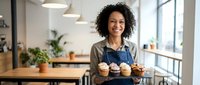 Young woman with curly dark hair and medium skin tone, wearing denim apron, holding tray of cupcakes in modern bakery kitchen with natural lighting and plants.