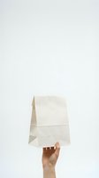 Hand holding blank white paper lunch bag against clean background for food delivery and takeaway packaging mockup.