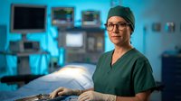 Female surgeon in green scrubs and surgical cap smiling confidently in modern operating room with medical equipment and monitors in background.