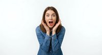 Young Caucasian woman in denim shirt showing shocked surprised expression with hands on cheeks and open mouth on white background isolated.