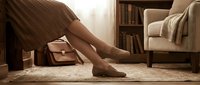 Barefoot woman in brown dress standing on beige carpet in cozy living room with armchair, books, and warm natural light for home decor and lifestyle content.