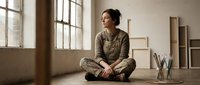 Young Asian woman in casual olive green clothing sitting cross-legged on wooden floor in bright minimalist studio space with large windows and natural lighting.