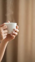 Woman hands holding steaming white coffee cup with neutral manicure against warm beige background for cozy morning lifestyle and relaxation concepts.