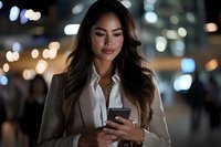 Young Asian businesswoman checking smartphone at night with city bokeh lights background, wearing beige blazer and white blouse, professional look.