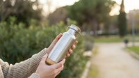 Hand holding white stainless steel water bottle in park setting with blurred green trees and pathway background for healthy lifestyle and hydration concept.