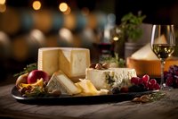 Gourmet cheese board with assorted artisanal cheeses, fresh fruits, grapes, and nuts served with white wine on rustic wooden table against blurred restaurant background.