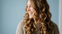 Young Caucasian woman with curly blonde hair smiling in cozy brown sweater against soft blue background for lifestyle and fashion content.