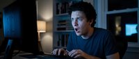 Young man with curly dark hair and olive complexion, wearing dark t-shirt, working late at computer in home office with blue lighting, focused on screen with keyboard and mouse for tech work.