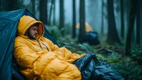 Young man in yellow raincoat resting inside blue tent during rainy weather in misty pine forest. Atmospheric outdoor camping scene with moody dark tones.