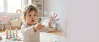 Adorable toddler girl with curly hair reaching out with open hand in bright modern nursery room with natural sunlight and colorful toys in background.