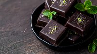 Dark chocolate squares with gold dust and fresh mint leaves arranged on black plate against dark background. Luxury dessert photography for culinary use.