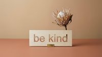Wooden sign with be kind inscription and dried protea flower on beige background, minimalist decor for kindness and wellness concepts.