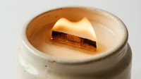 Burning wooden wick candle in a rustic ceramic jar with warm glowing flame, cozy home decor and aromatherapy concept.