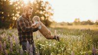 Loving father playing with toddler son in wildflower meadow during golden hour sunset, family bonding outdoors in nature for parenting concepts.