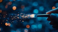 Magical glowing sparkles trail from magic wand against dark blue bokeh background, creating ethereal light effect with orange and gold particles.