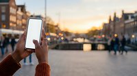 Close-up of hand holding smartphone with blank white screen against urban sunset backdrop, pedestrians and historic architecture blurred in background for app mockup.