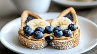 Playful cat-shaped toast with peanut butter, fresh banana slices and blueberries on white plate. Powdered sugar dusting adds whimsical breakfast appeal.