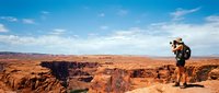 Young male hiker with backpack standing on rocky desert plateau, photographing vast canyon landscape under blue sky with clouds.
