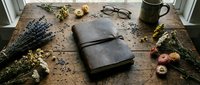 Vintage leather journal on rustic wooden table surrounded by dried wildflowers, lavender, and antique glasses, evoking nostalgia and creativity.