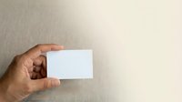 Male hand with medium-deep skin tone holding blank white business card against neutral beige wall background. Suitable for corporate branding and business identity mockups.
