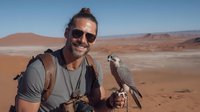 Smiling male traveler with sunglasses holding falcon in desert landscape with red sand dunes and mountains in background.