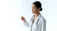 Young female healthcare professional of East Asian descent wearing white medical coat with stethoscope, gesturing while explaining or presenting medical information against neutral background.