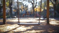 Empty swing in sunlit playground with blurred autumn background creates nostalgic mood. Natural bokeh effect and golden hour lighting evoke childhood memories.