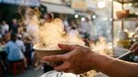 Man's hand holding steaming bowl of food at crowded street market or festival, steam rising from hot meal, street food vendor serving traditional cuisine.