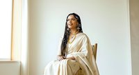 Young woman with dark braided hair and gold jewelry, wearing a cream-colored draped garment, seated indoors by natural light. Suitable for lifestyle, fashion, and portrait projects.