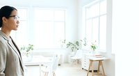 Young Asian woman in business casual attire standing in bright, minimalist office space with natural light, potted plants, and modern furniture for corporate, professional, and workplace concepts.