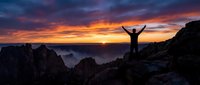 Silhouette of triumphant hiker with raised arms celebrating success on mountain peak during spectacular sunrise with dramatic orange and purple sky.