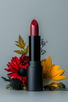 Red matte lipstick in black tube with autumn flowers arrangement featuring red gerbera and yellow sunflower on gray background, beauty product photography.