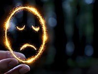 Glowing sparkler drawing sad face emoji in dark background with bokeh lights, expressing negative emotions and melancholy mood through light painting technique.