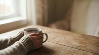 Cozy morning scene with hands in knit sweater holding warm coffee mug on rustic wooden table by window with soft natural light for lifestyle content.