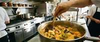 Chef stirring large pot of paella with seafood and saffron rice in professional kitchen with staff working in background.