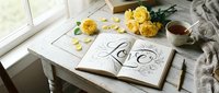 Open journal with hand-lettered Love calligraphy, yellow roses, and tea cup on rustic white wooden table by sunny window, cozy romantic mood.