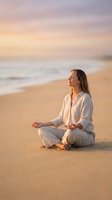 Peaceful woman meditating in lotus position on sandy beach during golden hour sunset with soft pastel sky and gentle ocean waves in background.