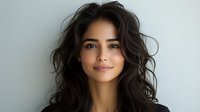 Young Hispanic woman with long dark wavy hair and natural makeup smiling at camera against light gray background, professional headshot style portrait.
