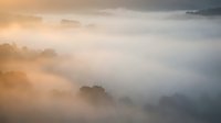 Misty mountain landscape with golden sunrise light breaking through fog and clouds over forested hills creating atmospheric layers and dramatic mood.