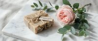 Handmade soap bar wrapped in kraft paper with twine on marble surface, styled with fresh pink rose and eucalyptus greenery for natural skincare, wellness and spa product marketing.