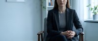 Professional businesswoman in dark suit sitting confidently in modern office environment with hands clasped, representing corporate leadership and workplace success.