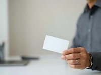 Young professional man of mixed ethnicity wearing gray shirt holding blank white business card at office desk for corporate branding and networking purposes.
