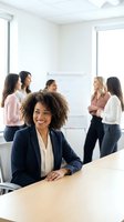 Confident African American businesswoman smiling at camera during team meeting in modern office with diverse female colleagues collaborating in background.