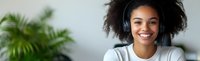 Young African American woman with natural curly hair wearing headphones and white t-shirt smiling broadly against blurred background with green plant.